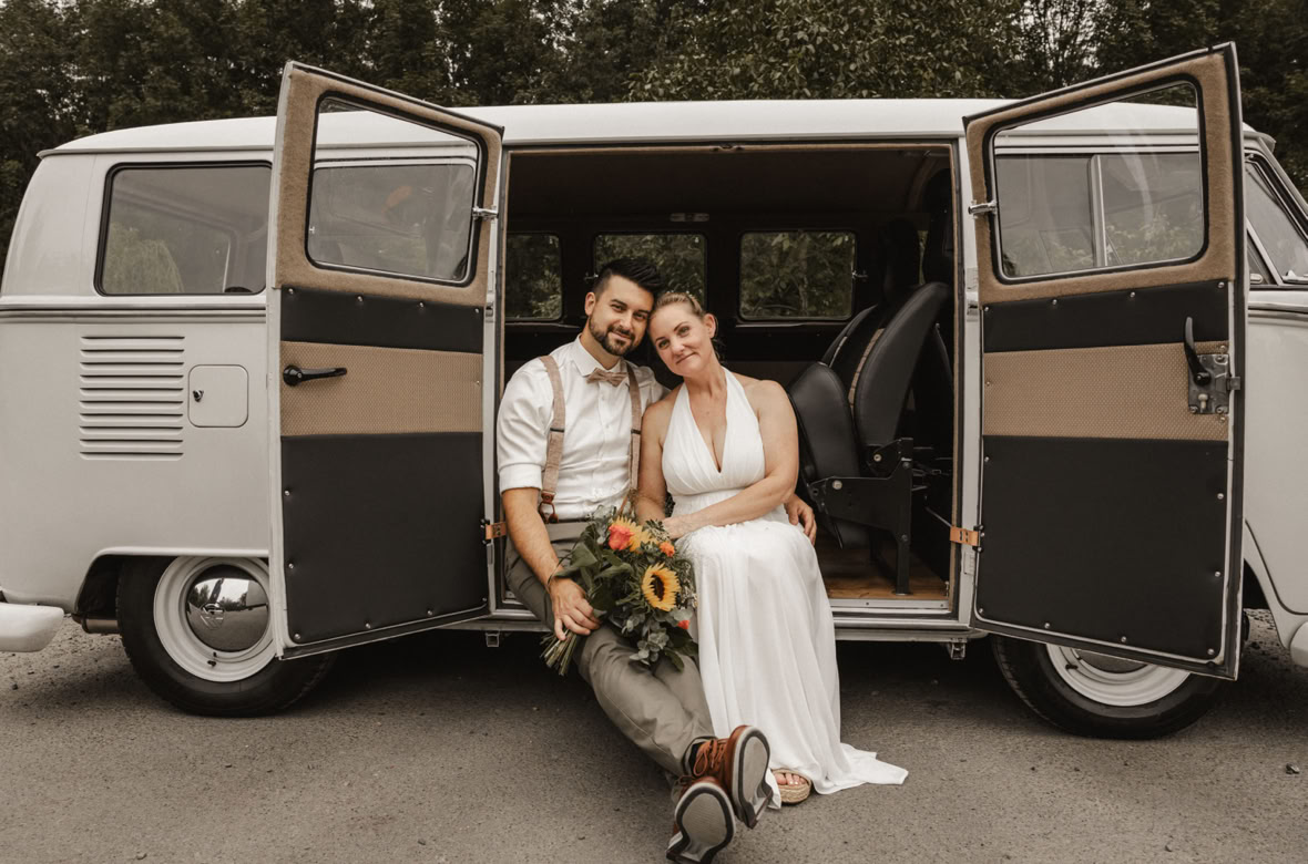 Ein Paar sitzt lächelnd vor einem geöffneten, weißen VW-Bus. Die Frau hält einen Blumenstrauß.