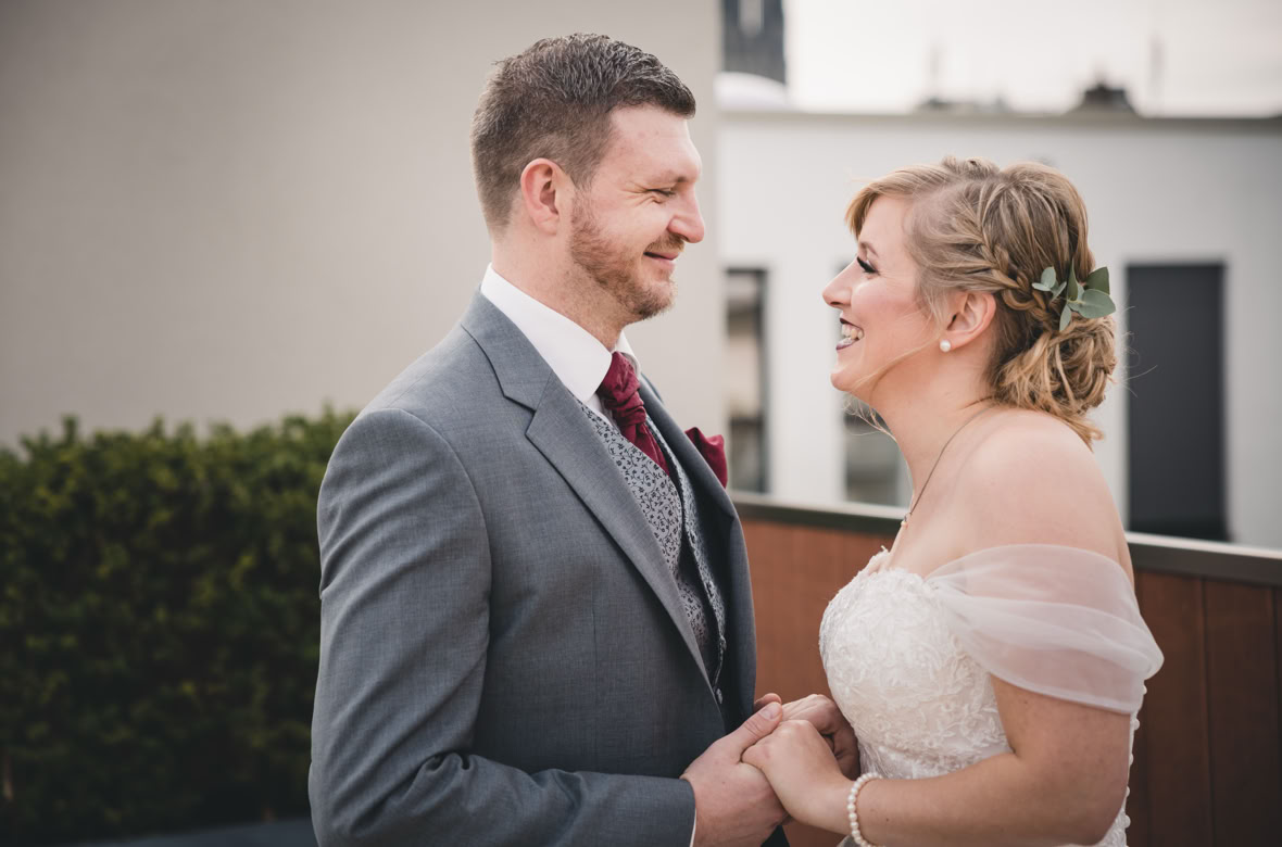 Ein lächelndes Paar in Hochzeitskleidung hält sich an den Händen und steht sich gegenüber, im Hintergrund sind Gebäude und Pflanzen zu sehen.